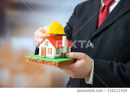 A businessman places a yellow construction hard hat on his house. Housing renovation and reconstruction. Affordable housing and infrastructure. Urban development improves living standards and supports 138121329