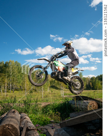 Motocross rider jumping on motorcycle over log obstacle. Professional man performing extreme stunt in open field. Competitive dirt bike racing skill, adventure and active outdoor sport. Motocross rider jumping on motorcycle over log obstacle. Professional man performing extreme stunt in open field. Competitive dirt bike racing skill, adventure and active outdoor sport. 138121562