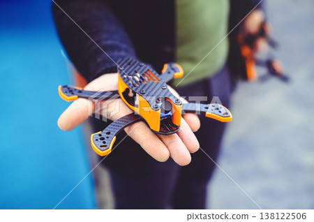 A horizontal close-up shot of a person holding a compact FPV racing drone in their hand. The drone features a black carbon fiber frame with bright orange 3D-printed accents and a blue camera lens. A horizontal close-up shot of a person holding a compact FPV racing drone in their hand. The drone features a black carbon fiber frame with bright orange 3D-printed accents and a blue camera lens. 138122506