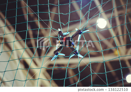 A horizontal shot of a small FPV racing drone with blue propellers stuck in a green safety net during an indoor flight. A horizontal shot of a small FPV racing drone with blue propellers stuck in a green safety net during an indoor flight. 138122510