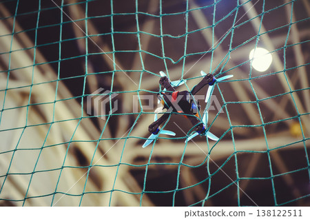 A horizontal shot of a small FPV racing drone with blue propellers stuck in a green safety net during an indoor flight. A horizontal shot of a small FPV racing drone with blue propellers stuck in a green safety net during an indoor flight. 138122511