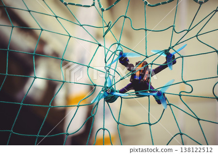 A horizontal shot of a small FPV racing drone with blue propellers stuck in a green safety net during an indoor flight. 138122512