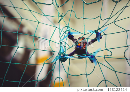 A horizontal shot of a small FPV racing drone with blue propellers stuck in a green safety net during an indoor flight. A horizontal shot of a small FPV racing drone with blue propellers stuck in a green safety net during an indoor flight. 138122514