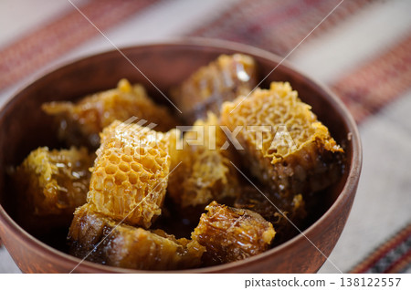 A close-up horizontal shot of golden, fresh honeycombs served in a brown clay bowl. The bowl sits on a white tablecloth with traditional red embroidered patterns. A close-up horizontal shot of golden, fresh honeycombs served in a brown clay bowl. The bowl sits on a white tablecloth with traditional red embroidered patterns. 138122557