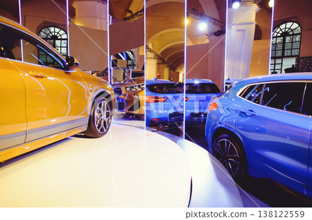 A wide horizontal shot of a car exhibition featuring golden and blue luxury vehicles. The composition is framed by vertical neon light pillars, creating a futuristic atmosphere in an arched hall. A wide horizontal shot of a car exhibition featuring golden and blue luxury vehicles. The composition is framed by vertical neon light pillars, creating a futuristic atmosphere in an arched hall. 138122559