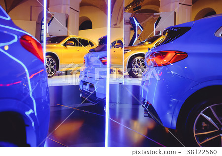 A wide-angle horizontal shot of a luxury car dealership, featuring a row of premium golden and blue vehicles parked under a series of illuminated arches and neon vertical pillars. A wide-angle horizontal shot of a luxury car dealership, featuring a row of premium golden and blue vehicles parked under a series of illuminated arches and neon vertical pillars. 138122560