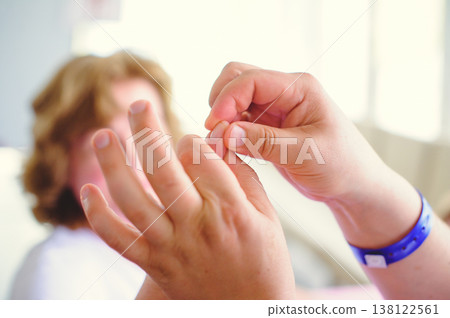 A horizontal close-up shot of a specialist's hands carefully examining or massaging a patient's finger. The background shows a blurred person in a bright clinic setting, and the specialist is wearing 138122561