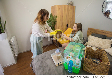 In a cozy bedroom, two girls are focused on their activity. One girl is handing a yellow cloth In a cozy bedroom, two girls are focused on their activity. One girl is handing a yellow cloth 138122593