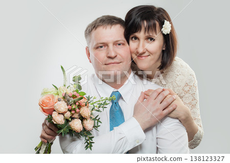 A woman holds a bouquet while hugging a man from behind in a studio. Both smile and look happy. A woman holds a bouquet while hugging a man from behind in a studio. Both smile and look happy. 138123327