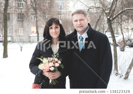 Couple standing together in winter street holding flowers in snowy park near buildings on a cold day Couple standing together in winter street holding flowers in snowy park near buildings on a cold day 138123329