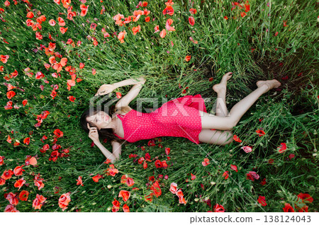 A horizontal high-angle shot of a young woman with long dark hair, wearing a red patterned summer dress, lying gracefully among green grass and vibrant blooming red poppies 138124043