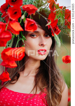 A vertical  portrait of a beautiful young woman with long dark hair and red lipstick, wearing a red patterned dress. Her face is artfully framed by a large wreath of bright red poppies, as she 138124085