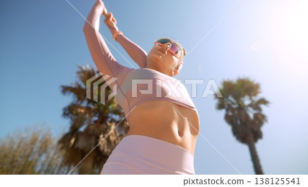 Woman in pink top and white shorts exercising beneath palm trees 138125541