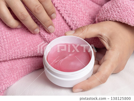 Woman in pink bathrobe holding white jar with eye patches in hands over white bedsheet Woman in pink bathrobe holding white jar with eye patches in hands over white bedsheet 138125800