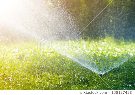 Lawn water sprinkler spraying water over lawn green fresh grass in garden or backyard on hot summer day. Automatic watering equipment, lawn maintenance, gardening and tools concept. 138127438
