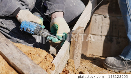 Construction Worker Drilling Wood Form Near Foundation, Kneeling In Gloves And Boots, Securing Boards With Screws, Earth And Tools Visible, Handson Framing Task During Daytime Build Construction Worker Drilling Wood Form Near Foundation, Kneeling In Gloves And Boots, Securing Boards With Screws, Earth And Tools Visible, Handson Framing Task During Daytime Build 138128903