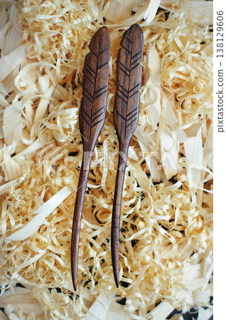 Vertical close-up of two handmade wooden hair sticks shaped like feathers, resting in a bed of wood shavings. 138129606