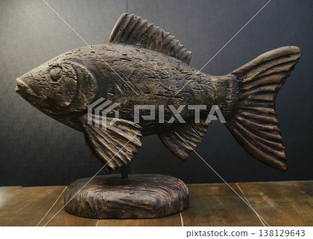 Hand-carved wooden fish sculpture with aged, weathered texture on a solid timber stand against a dark background. Hand-carved wooden fish sculpture with aged, weathered texture on a solid timber stand against a dark background. 138129643