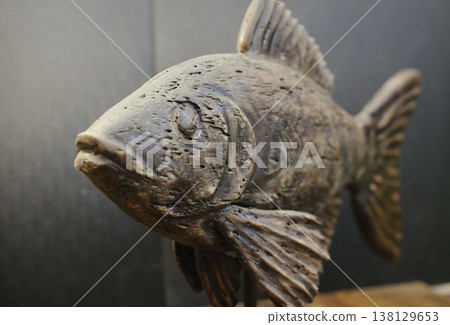 Close-up perspective view of a hand-carved wooden fish sculpture, highlighting the detailed weathered head and textured fins on a dark background. Close-up perspective view of a hand-carved wooden fish sculpture, highlighting the detailed weathered head and textured fins on a dark background. 138129653