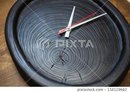 Close-up perspective of a handcrafted wall clock made from a scorched black wood slice, emphasizing the rhythmic circular annual rings and silver hands. Close-up perspective of a handcrafted wall clock made from a scorched black wood slice, emphasizing the rhythmic circular annual rings and silver hands. 138129662