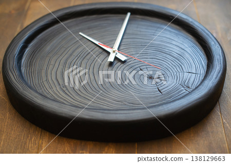 High-angle perspective of a handcrafted wall clock featuring a scorched black wood slice with deep annual ring patterns and minimalist silver hands. High-angle perspective of a handcrafted wall clock featuring a scorched black wood slice with deep annual ring patterns and minimalist silver hands. 138129663