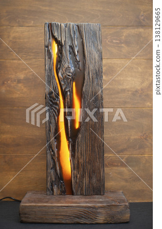 Handcrafted decorative table lamp made from a weathered solid wood slab with an internal warm orange glow, highlighting the natural textures and cracks. 138129665