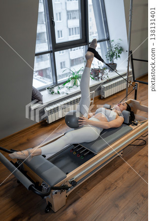 Fit woman stretching legs on pilates reformer by sunny window in modern indoor pilates studio. Stretching, pilates reformer, fitness, sunny, studio. Fit woman stretching legs on pilates reformer by sunny window in modern indoor pilates studio. Stretching, pilates reformer, fitness, sunny, studio. 138131401