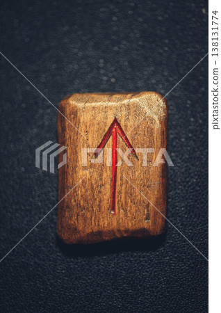 A vertical close-up of a handcrafted wooden Teiwaz rune stone, featuring the carved arrow symbol filled with red pigment on a rustic grain. 138131774