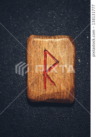 A vertical close-up of a handcrafted wooden Raido rune stone, featuring the carved symbol filled with red pigment on a rustic grain. A vertical close-up of a handcrafted wooden Raido rune stone, featuring the carved symbol filled with red pigment on a rustic grain. 138131777