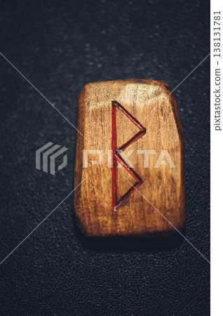 A vertical close-up of a handcrafted wooden Berkana rune stone, featuring the carved B-shaped symbol filled with red pigment on a rustic grain. 138131781