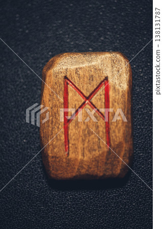Vertical close-up of a handcrafted wooden Mannaz rune stone, featuring the carved symbol filled with red pigment on a rustic grain. 138131787