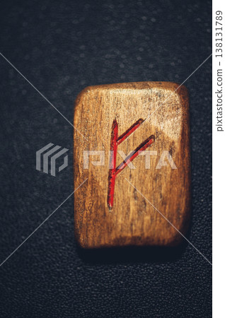 Vertical close-up of a handcrafted wooden Fehu rune stone, with the carved wealth symbol filled with red pigment on a rustic grain. 138131789