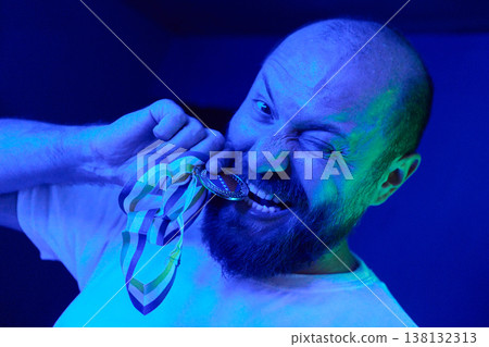 Bearded Man Biting Medal in Blue Light Celebrating Victory and Playful Achievement Portrait 138132313