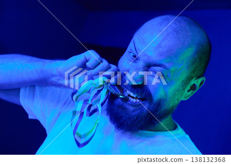Triumphant Man Biting Medal in Blue Light Celebrating Victory and Achievement With Playful Expression Triumphant Man Biting Medal in Blue Light Celebrating Victory and Achievement With Playful Expression 138132368