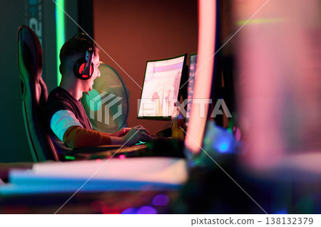 Focused Gamer Streaming Competitive PC Gameplay in Neon Lit Gaming Room With Headset and Monitor Focused Gamer Streaming Competitive PC Gameplay in Neon Lit Gaming Room With Headset and Monitor 138132379