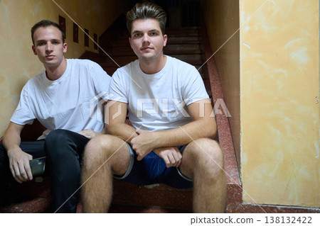 Two Young Men Sitting on Stairs in Casual White Tees Relaxing and Posing Together Two Young Men Sitting on Stairs in Casual White Tees Relaxing and Posing Together 138132422