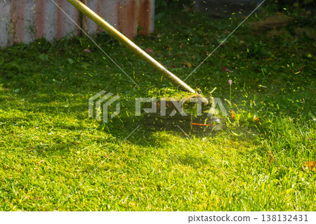 Grass trimmer cutting lawn close up. Line trimmer in action mowing green grass. Gardening equipment for yard maintenance and landscaping work. Power tool for lawn care at sunny summer day. Grass trimmer cutting lawn close up. Line trimmer in action mowing green grass. Gardening equipment for yard maintenance and landscaping work. Power tool for lawn care at sunny summer day. 138132431