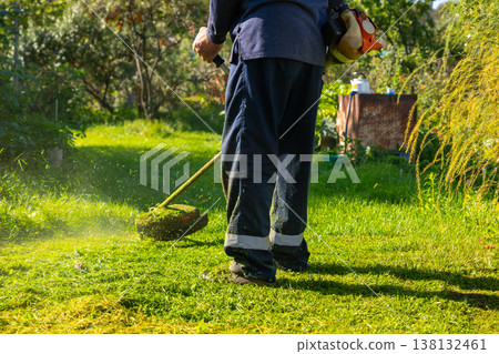 Gardener trimming grass in backyard. Man using string trimmer for lawn maintenance. Professional landscaping service for home garden improvement, summer yard work and outdoor lawn care. Gardener trimming grass in backyard. Man using string trimmer for lawn maintenance. Professional landscaping service for home garden improvement, summer yard work and outdoor lawn care. 138132461