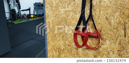 Suspension training straps hanging on a gym wall with fitness equipment in the background. Concepts of functional training, fitness lifestyle, and modern workout routines. Suspension training straps hanging on a gym wall with fitness equipment in the background. Concepts of functional training, fitness lifestyle, and modern workout routines. 138132474
