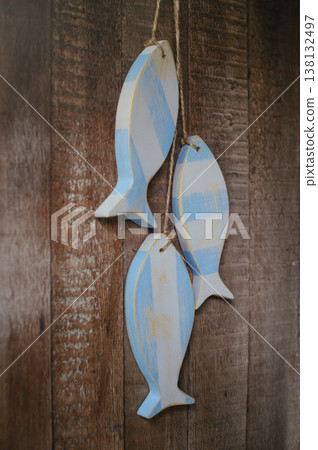 A vertical view of three handcrafted wooden fish sculptures with a distressed light blue and white stripe pattern, hanging on twine against a rustic wooden plank wall. 138132497