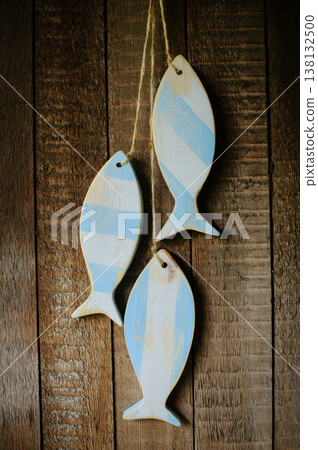 A vertical view of three handcrafted wooden fish sculptures with a distressed light blue and white stripe pattern, hanging on twine against a rustic wooden plank wall. 138132500