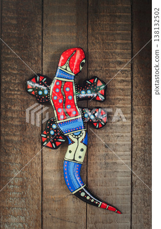Vertical close-up of a handcrafted, brightly painted wooden gecko lizard sculpture with folk patterns, mounted on weathered rustic planks. 138132502