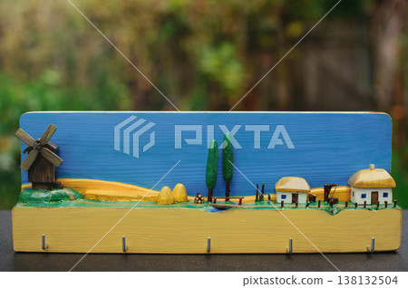 A horizontal close-up of a handcrafted wooden wall-mounted key holder, featuring a brightly painted miniature Ukrainian village diorama. A horizontal close-up of a handcrafted wooden wall-mounted key holder, featuring a brightly painted miniature Ukrainian village diorama. 138132504