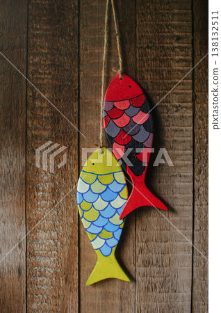 Vertical close-up of two handcrafted wooden fish sculptures, one yellow and one red with painted scales, hanging by twine against a rustic wooden wall. Vertical close-up of two handcrafted wooden fish sculptures, one yellow and one red with painted scales, hanging by twine against a rustic wooden wall. 138132511