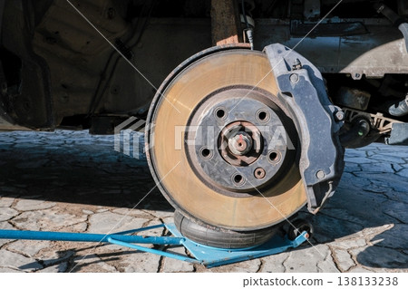 A car brake disc with visible rust and dust, mounted on a vehicle lifted by a jack. Brake caliper 138133238