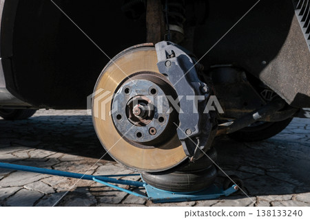 A car brake disc with visible rust and dust, mounted on a vehicle lifted by a jack. Brake caliper 138133240