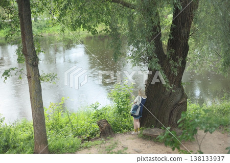 Kid with backpack gazes at shimmering water beneath leafy canopy Kid with backpack gazes at shimmering water beneath leafy canopy 138133937