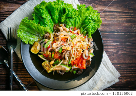 Delicious papaya salad or Tum Thai with squid tentacles, Lentils, nuts, raw papaya, fish sauce, sugar, tomato, and lime. Cutlery is placed on the wooden floor. Taken in a photo studio. Thai food. 138136062