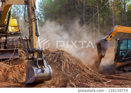 Two pieces of heavy machinery work to move dirt clear ground on construction site surrounded by trees. 138138754