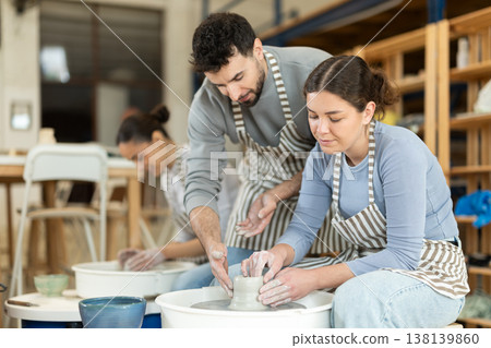 Girl craftsperson busy molding clay products using pottery wheel in lesson with male teacher Girl craftsperson busy molding clay products using pottery wheel in lesson with male teacher 138139860